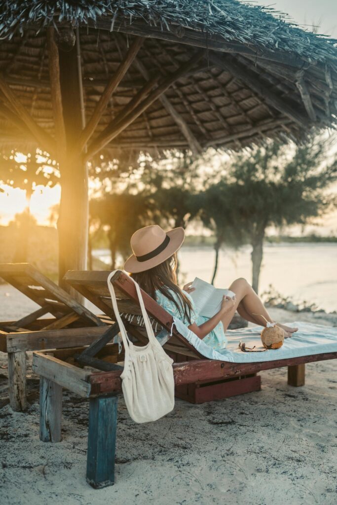 A woman reads by the beach under a thatch umbrella in Zanzibar at sunset.